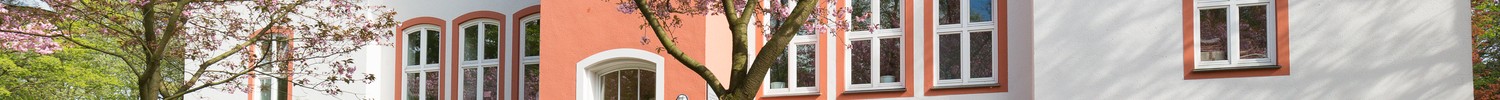 Weißes, zweistöckiges Schulgebäude mit rotem Dach, im Vordergrund grüner Rasen und rosa blühende Kirschbäume.