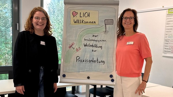 Zwei Frauen stehen neben einer Infotafel mit Informationen für die Weiterbildung zur PraxisanleiterIn
